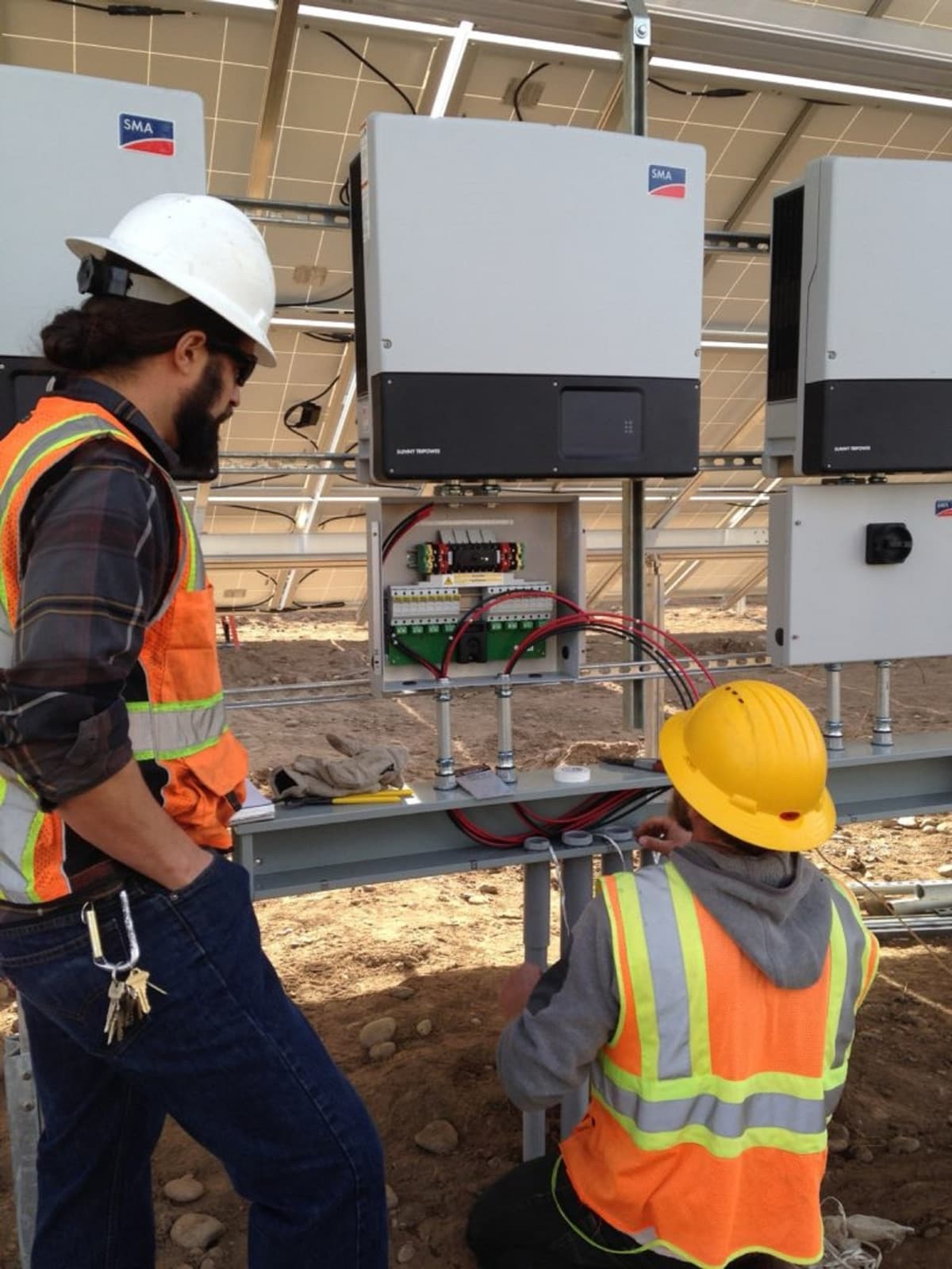 Technicians working at wall-mounted string inverters with an open combiner box, DC string conductors visible